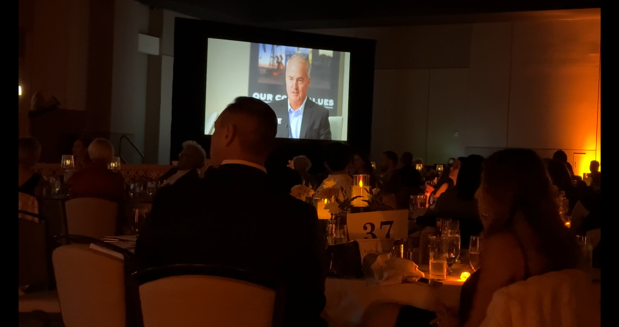 Corporate Sponsorship video playing on a large screen in front of an audience in a darkened room.